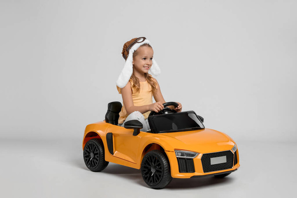 Cute little girl in pilot hat driving children's electric toy car on grey background - Fotografie, Obrázek