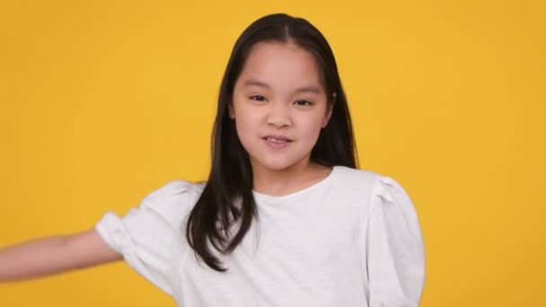 Engraçado pouco asiático menina mostrando bíceps, demonstrando minúsculos músculos, sorrindo para câmera sobre laranja estúdio fundo - Filmagem, Vídeo