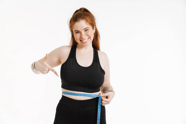 Young ginger woman with measuring tape pointing finger at her waist isolated over white background - Photo, image