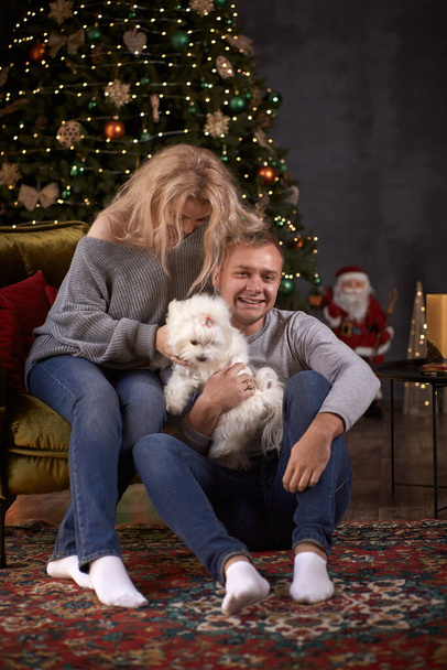 Pareja romántica con su perro se burlan cerca del árbol de Navidad en el interior de la casa acogedor estético festivo. Buen humor de la historia de amor de Navidad. Candid, verdadero momento - Foto, imagen