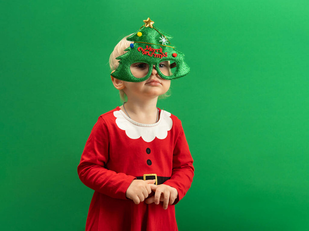  Happy Toddler girl in red Christmas costume and green tree glasses smiling on green background COPYSPACE - Фото, изображение