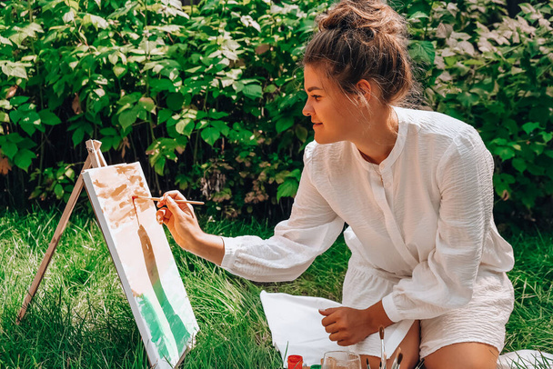 Художник рисует на мольберте на открытом воздухе в саду. Открытая художественная мастерская. Нарисуйте холст кистью и палитрой, сидя на траве во время пикника в парке - Фото, изображение