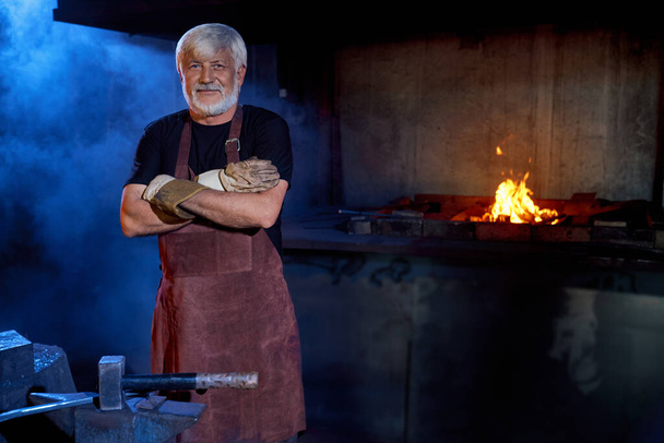 Aged blacksmith in safety apron and gloves posing at forge - Foto, immagini