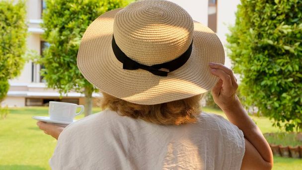 Une femme heureuse dans un chapeau de paille tient une tasse de boisson chaude et boit du café, assise le matin sur une terrasse d'été par une journée ensoleillée. Bonnes manières en buvant du thé. Le concept de bonjour, voyage - Photo, image