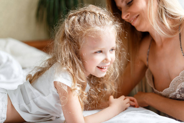 Portrait of daughter and mother spend morning together at home. Beautiful child girl Caucasian kid with mom in bed. Loving caring mom makes child wake up - Fotó, kép