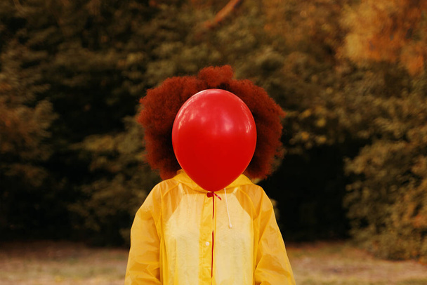 Sarı yağmurluklu bir kadın ormanın arka planında kırmızı bir balon tutuyor.. - Fotoğraf, Görsel