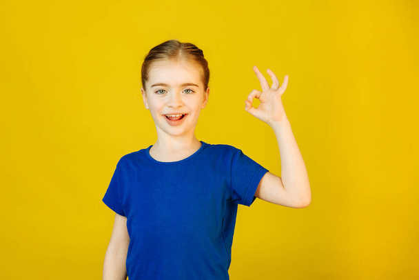 Little girl looking at camera and showing OK gesture in sign language on yellow background. - Valokuva, kuva