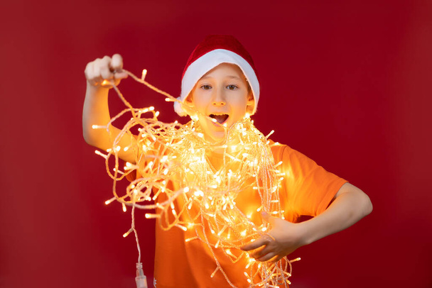 menino feliz em um chapéu de Papai Noel vermelho, enredado em uma grinalda de Natal que brilha - Foto, Imagem