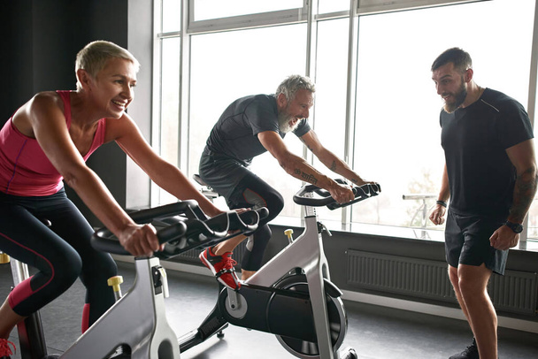 Young sportive instructor watching couple cycling in gym - Photo, image