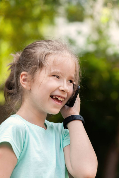 Gelukkige vrolijke basisschool leeftijd kind, meisje praten op haar mobiele telefoon, met een smartphone naast het oor dragen van een smartwatch, lachen. Buitenportret, gezichtsclose-up, communicatie concept - Foto, afbeelding