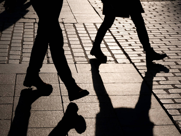 Δύο ανώνυμοι αγνώριστοι άνθρωποι περπατούν στο δρόμο, σκούρες μαύρες σιλουέτες ποδιών, σκιές. Πεζοί σε εξωτερικούς χώρους, κοινωνία, ανωνυμία, μοναξιά και θλίψη, κοινωνικά θέματα αφηρημένη έννοια - Φωτογραφία, εικόνα