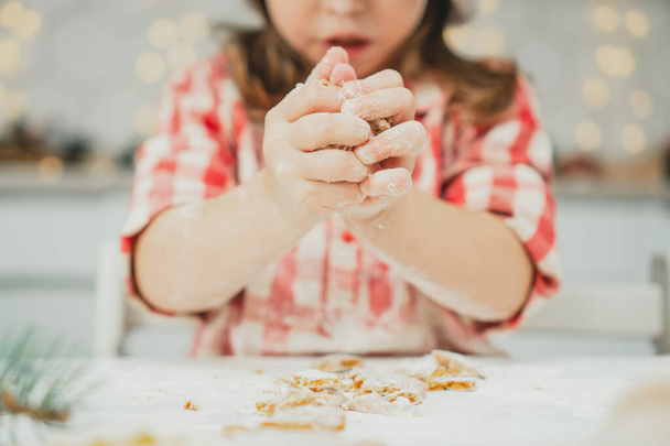 Partially blurred hands of little girl in red checkered shirt prepare dough for gingerbread cookies in white kitchen. Child knead dough on white table strewn with flour - Photo, image