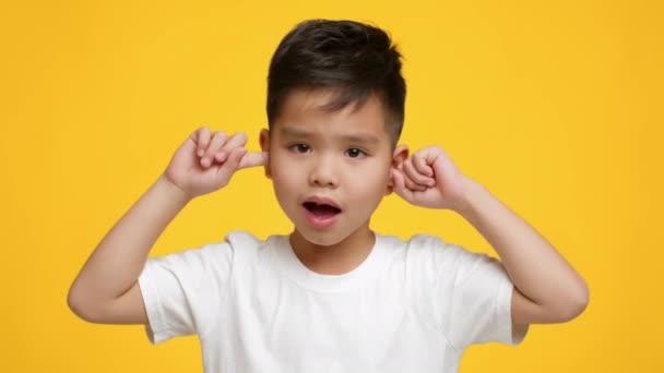 ondeugende aziatische kleine jongen bedekken oren schudden hoofd, gele achtergrond - Video