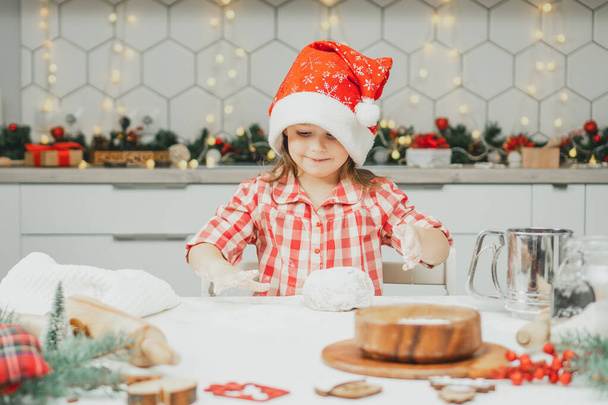 Little dark-haired girl 3 years old in red Christmas cap and checkered shirt prepares dough for gingerbread cookies in white Christmas decorated kitchen with garland lights. Merry Christmas, New Year - Photo, Image