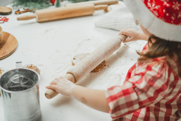 Mani parzialmente sfocate di bambina in berretto rosso di Natale e camicia a quadretti stendono la pasta per biscotti di pan di zenzero con mattarello in cucina bianca. Il bambino prepara la pasta su tavolo bianco con farina - Foto, immagini