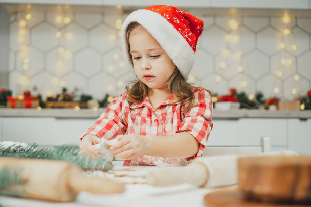 Kis sötét hajú lány 3 éves piros karácsonyi sapkában és kockás ingben kivágja mézeskalács sütik hengerelt tészta fehér karácsonyi díszített konyha. Boldog karácsonyt, boldog új évet! - Fotó, kép
