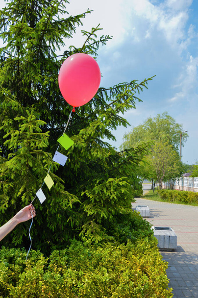 Una mano femenina sostiene un globo rojo sobre el fondo de un abeto verde. Hay pegatinas de papel remachadas a la cinta de la bola, en las que se escriben los deseos. - Foto, Imagen