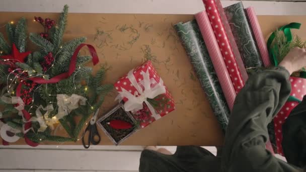 Plan odgórny. W pełni widoczny stół z dekoracjami. Kobiece ręce umieścić i sfinalizować prezent świąteczny zawinięty w papier rzemieślniczy na drewnianym stole. Taśma bandażowa  - Materiał filmowy, wideo
