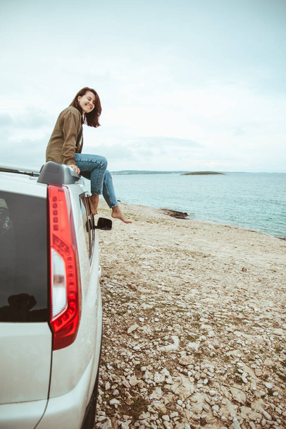 車の屋根の上に座って海の景色を楽しんでいる女性。夏休みロードトリップ - 写真・画像