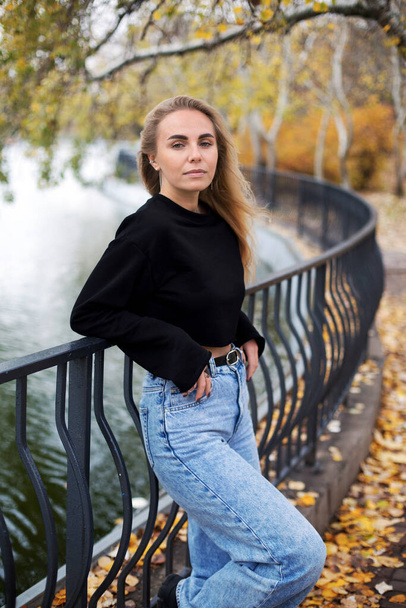 belle femme blonde caucasienne en pull noir jeans marchant près d'un étang dans le parc d'automne - Photo, image