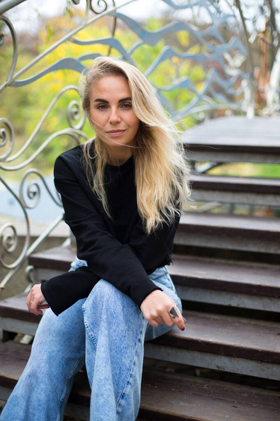 mooie Kaukasische blonde vrouw in een zwarte trui en jeans zit op de brugtrap in het najaarspark - Foto, afbeelding