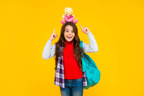 smiling teen girl in casual clothes holding school bag and toy bear on yellow background, school - Fotó, kép
