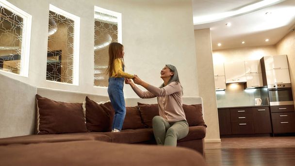 Grandmother supporting jumping granddaughter while sitting on sofa - Photo, image