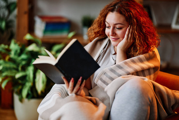 Ein schönes Ingwermädchen sitzt in eine Decke gehüllt im Wohnzimmer und liest morgens ein interessantes Buch. - Foto, Bild