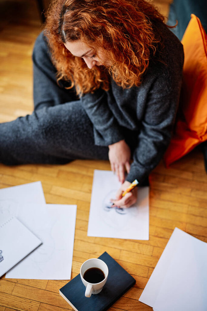 The top view of an artistic ginger girl is sitting on the floor in the morning and drawing portraits. She is enjoying her morning moments with a cup of fresh coffee. - Photo, Image