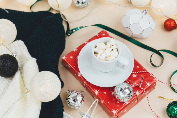 Cup of coffee, gift box and knitted sweaters, led lights on beige background. Hygge style, winter concept. Cozy home desk. New year and Christmas celebration mockup. Christmas still life - 写真・画像