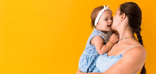Motherhood, hot summer - Banner young pregnant happy smiling mother woman in blue dress holding little baby daughter toddler sibling in arms looking into eyes kisses on yellow background coppy space - Foto, Bild