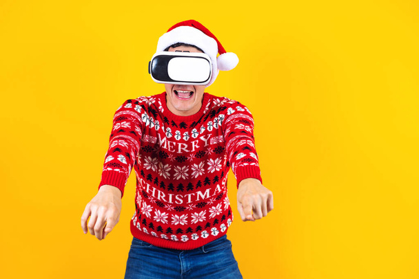 νεαρός Λατίνος gamer άνθρωπος στην εικονική πραγματικότητα headset gadget φορώντας Σάντα καπέλο και χριστουγεννιάτικο πουλόβερ σε κίτρινο φόντο. Χριστούγεννα και το χειμώνα vr έννοια της τεχνολογίας στο Μεξικό Λατινική Αμερική - Φωτογραφία, εικόνα