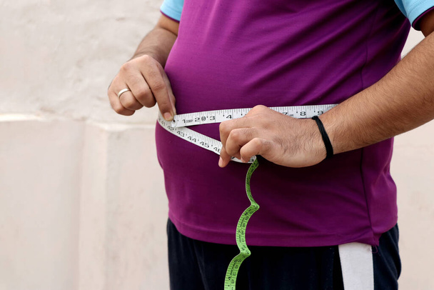 Un asiático mide su vientre gordo con una cinta métrica sobre un fondo liso - Foto, Imagen