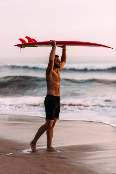 Male surfer on the beach at sunset. A sporty man with a surfboard. The guy is a surfer. An attractive young guy is standing on the beach with a red surfboard in his hands. Copy space - Fotó, kép
