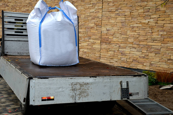 ładowanie dużych białych plastikowych toreb pełnych pokryć dachowych na mniejszą ciężarówkę. dźwig trzymający w powietrzu metr sześcienny ciężkiego żwiru za uchwyty na haku. - Zdjęcie, obraz