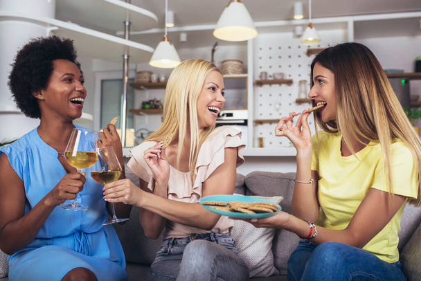 Gelukkig jonge vrouwelijke vrienden met wijn en koekjes op de bank thuis.  - Foto, afbeelding
