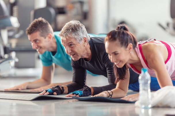 Group of people in various age and gender having fun while doing group exercise of elbow plank in the gym.  - Valokuva, kuva