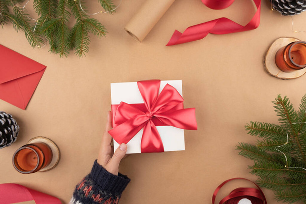 Weiße Geschenkschachtel mit roter Schleife hält in der Hand auf weißem Hintergrund. Verpackung und Vorbereitung von Geschenken für die Feier. Weihnachten Silvesterkulisse mit Baumzweigdekor und Lichtern - Foto, Bild