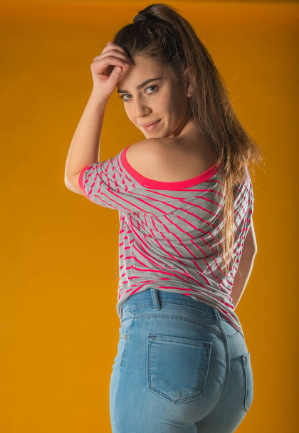 Beautiful young woman in pink mini dress posing with hand on hip, presenting something and looking away. Three quarter length studio shot on yellow background. - Photo, Image