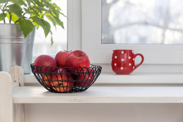 Red apples in a black mesh basket are on a white wooden shelf. There is a red cup of tea with white polka dots on the windowsill. Space for text. Copy space - Фото, зображення