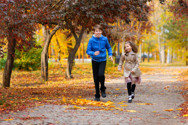 niños corren a lo largo del camino en el parque de otoño. juegan y ríen, se divierten mucho. están rodeados de hermosa naturaleza y árboles con hojas amarillas y rojas - Foto, Imagen
