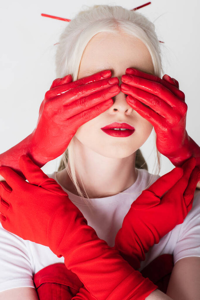 Female hands in red paint covering eyes of albino model isolated on white - Photo, Image
