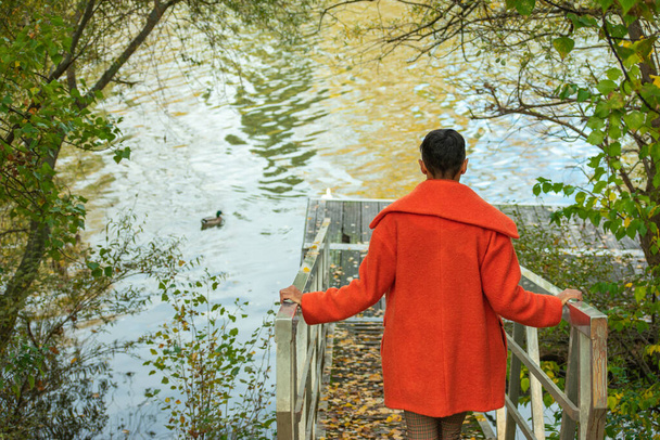 μια γυναίκα στην πλάτη της με πορτοκαλί παλτό πηγαίνει κάτω στην προβλήτα του ποταμού - Φωτογραφία, εικόνα