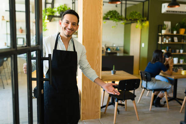 Saludando a los clientes. Joven guapo y dueño de un negocio sonriendo y abriendo la puerta a los clientes en su cafetería - Foto, Imagen