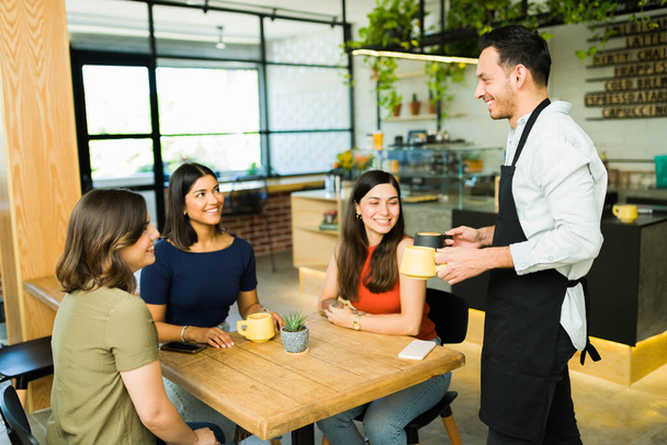 Happy ober brengt kopjes koffie naar de tafel van opgewonden vrouwelijke klanten in de coffeeshop - Foto, afbeelding