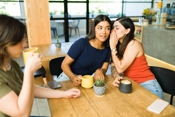 Jovem mulher sussurrando um segredo para seu melhor amigo enquanto sentado em um belo restaurante de café  - Foto, Imagem