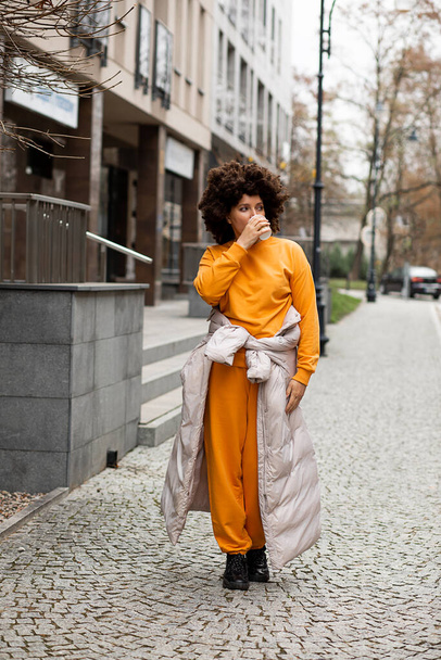 Portrait of Young, beautiful caucasian woman 40 years old, Sport casual clothes, walking on street, Disposable Cup of coffee. Afro Hairstyle. Enjoying life on autumn day. Cheerful people, City life - Foto, imagen