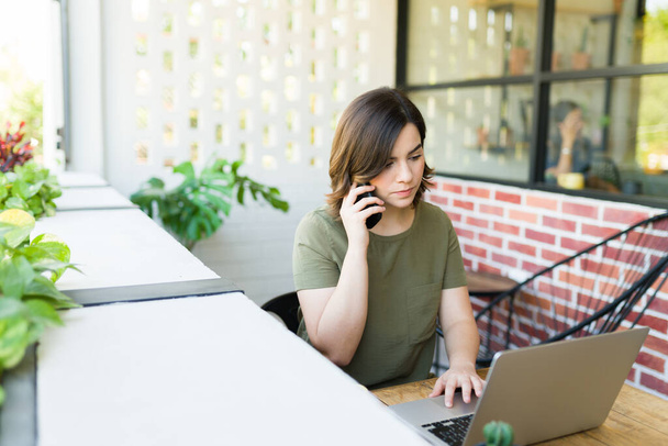 Многозадачная молодая женщина, делающая деловой звонок и сидящая на ноутбуке во дворе кофейни - Фото, изображение