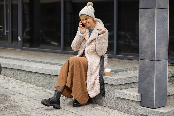 Mladá roztomilá žena s kávou v jednorázovém šálku, pomocí mobilního telefonu, sedí s úsměvem na schodech na evropské ulici, na sobě kožich faux kabát a klobouk, styl ulice, moderní život - Fotografie, Obrázek