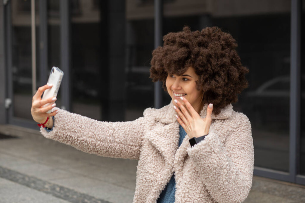 Young cute caucasian woman with Afro hairstyle wig making selfie or video call in city street on smartphone mobile phone. Influencer blogger create content for social media or blog. Lifestyle photo - Zdjęcie, obraz